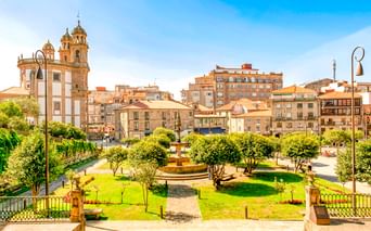 Plaza de la Herrería in Pontevedra mit grünem Park, Brunnen und historischer Kirche. Gebäude säumen den Platz unter blauem Himmel.