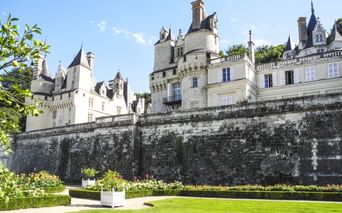 Schloss Ussé in Rigny-Ussé mit weißen Steinmauern, Türmchen und Türmen hinter einer verwitterten Terrassenmauer. Gepflegter Rasen und Gärten im Vordergrund.