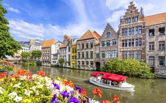 Weißes Touristenboot mit rotem Dach auf Kanal in Gent. Historische Treppengiebelhäuser säumen das Ufer, bunte Blumen im Vordergrund.