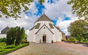 Weiße mittelalterliche Rundkirche Østerlars mit konischem Dach und Eingang, eingerahmt von grünen Bäumen unter blauem Himmel auf Bornholm.