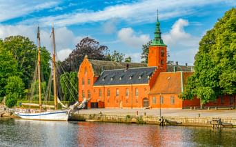 Orangefarbenes Schloss Halmstad mit Uhrturm am Wasser. Ein traditionelles Segelschiff liegt am Kai unter blauem Himmel mit weißen Wolken.