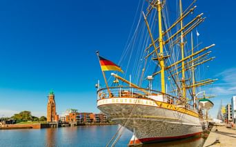 Weißes Segelschiff Deutschland mit deutscher Flagge am Neuen Hafen in Bremerhaven. Roter Backsteinleuchtturm und moderne Gebäude am Wasser.