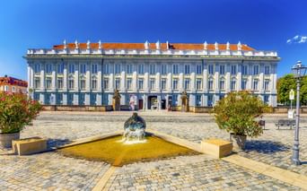 Residenz Ansbach, ein weißes Barockschloss mit orangefarbenem Dach, vom Kopfsteinpflasterplatz mit Brunnen und blühenden Pflanzen unter blauem Himmel.