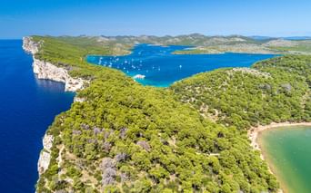 Luftaufnahme des Nationalparks Telascica mit türkisfarbener Bucht mit weißen Segelbooten, umgeben von grünen bewaldeten Klippen und felsiger Küste.