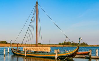 Wikingerschiff-Nachbau an einem Holzsteg auf ruhigem Wasser. Das Schiff hat einen hohen Mast, gebogenen Bug mit Drachenkopf und dunklen Rumpf mit gelber Verzierung.