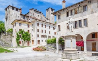 Historischer Platz in Feltre mit weißer Steinstatue von Vittorino da Feltre auf Sockel, umgeben von traditionellen italienischen Gebäuden mit Bögen.