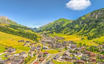 Luftaufnahme des Dorfes Lech am Arlberg in einem grünen Tal, umgeben von bewaldeten Bergen unter blauem Himmel mit vereinzelten Wolken.