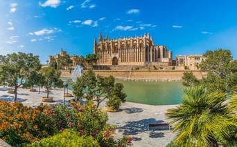 La Seu Kathedrale in Palma de Mallorca mit gotischer Architektur neben einem Teich, umgeben von bunten Gärten und Palmen unter blauem Himmel.
