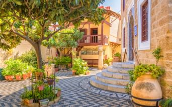 Sonniger Innenhof in der Altstadt von Rhodos mit gemustertem Steinpflaster, geschwungenen Stufen, Topfpflanzen, einem Baum und traditionellen Gebäuden.