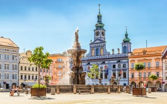 Historischer Brunnen mit Statue am Hauptplatz in Budweis, Tschechien. Blaues barockes Rathaus mit Glockenturm und bunte Gebäude umgeben den Platz.