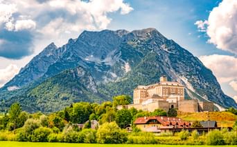 Schloss Trautenfels auf einem Hügel, umgeben von grünen Wiesen und Bäumen, mit einem markanten Berggipfel dahinter unter blauem Wolkenhimmel.