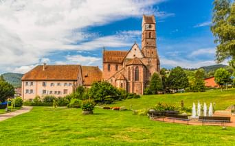 Kloster Alpirsbach mit rotem Sandsteinkirchturm und angrenzenden Gebäuden auf grüner Wiese mit Brunnen, umgeben von Bäumen und Hügeln.