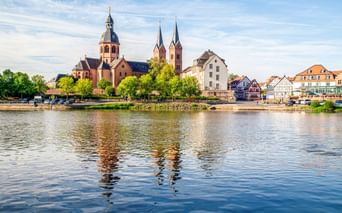 Blick über den Main auf Seligenstadt mit der Einhardsbasilika mit Zwillingstürmen und Kuppel, umgeben von historischen Gebäuden.