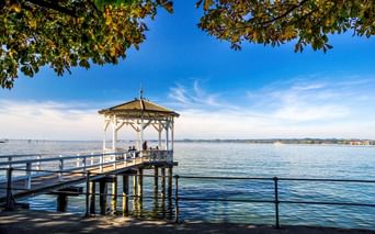 Holzsteg mit weißem Pavillon am Bodensee bei Bregenz. Menschen sitzen unter der überdachten Struktur, mit ruhigem blauen Wasser und ferner Küstenlinie.