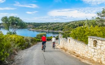 Zwei Radfahrer auf einer asphaltierten Straße neben einer Steinmauer mit blauer Bucht und grünen Hügeln der Insel Meganisi im Hintergrund.