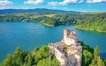 Luftaufnahme der Burg Niedzica auf einer bewaldeten Halbinsel, umgeben von türkisfarbenem Seewasser, mit Bergen und Wäldern im Hintergrund.