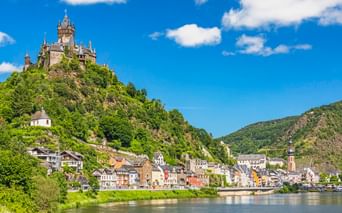 Stadt Cochem an der Mosel mit der Reichsburg auf einem grünen Hügel. Bunte Häuser säumen das Flussufer unter blauem Himmel.