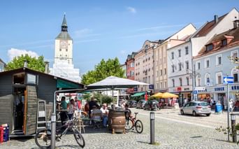 Kopfsteinpflaster-Marktplatz in Deggendorf mit weißem Uhrturm, bunten Gebäuden, Marktständen, geparkten Fahrrädern und Außensitzplätzen unter blauem Himmel.