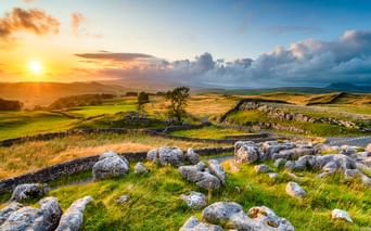 Weiße Kalksteine bei Winskill Stones mit grünen Feldern, Steinmauern und sanften Hügeln unter goldenem Sonnenuntergangshimmel.