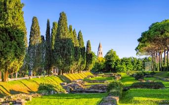Archäologische Stätte in Aquileia mit antiken Steinruinen auf grünen Rasenflächen, hohen Zypressen und dem Glockenturm der Basilika im Hintergrund.