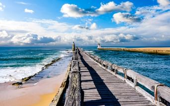 Holzsteg mit Geländer führt ins Meer in Capbreton, mit einem Leuchtturm am Ende und einer weißen Bake auf der rechten Mole.