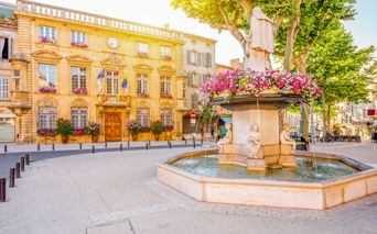 Steinbrunnen mit rosa Blumen in Salon-de-Provence. Gelbes historisches Gebäude mit Uhr und Flaggen im Hintergrund unter Bäumen.