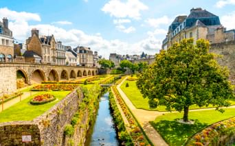 Formale Gärten mit bunten Blumenbeeten und Wasserkanal entlang mittelalterlicher Steinmauern und Bogenbrücke in Vannes, Frankreich.
