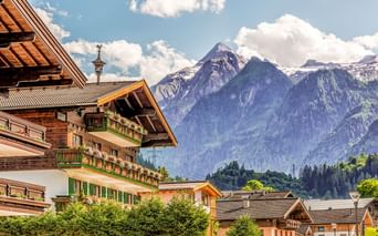 Traditionelle Alpenchalets in Kaprun mit blumengeschmückten Balkonen vor schneebedeckten Berggipfeln unter blauem Himmel mit weißen Wolken.