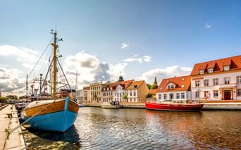 Harbor of Ueckermünde with blue wooden fishing boat in foreground and red boat behind. Historic buildings with orange-tiled roofs line the waterfront.