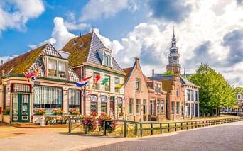 Traditionelle niederländische Gebäude mit bunten Flaggen entlang eines Kanals in Bolsward. Ein Kirchturm erhebt sich im Hintergrund unter blauem Himmel.