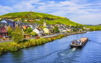 Frachtschiff auf der Mosel vor Wormeldange mit bunten Häusern, Kirchturm und weinbewachsenen Hügeln unter blauem Himmel.