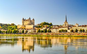 Schloss Saumur mit Türmen auf einem Hügel über der Stadt Saumur und der Loire. Das Schloss und die Gebäude spiegeln sich im Wasser.