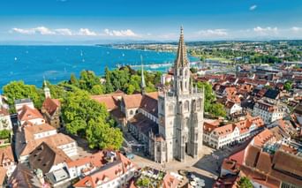 Luftaufnahme des Konstanzer Münsters mit gotischem Turm über den rotgedeckten Gebäuden. Bodensee mit Segelbooten im Hintergrund sichtbar.