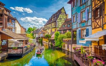 Bunte Fachwerkhäuser säumen einen Kanal in Klein-Venedig, Colmar. Ein Boot mit Touristen schwimmt auf dem grünen Wasser unter blauem Himmel.