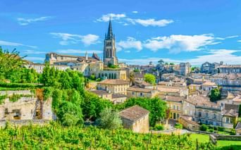 Panoramablick auf Saint-Émilion mit Steingebäuden, einem markanten Kirchturm und üppiger grüner Vegetation unter blauem Himmel mit weißen Wolken.
