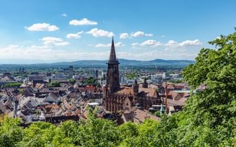Panoramablick auf die Freiburger Altstadt mit dem markanten Münsterturm über rotgedeckten Gebäuden, umgeben von grünen Hügeln unter blauem Himmel.