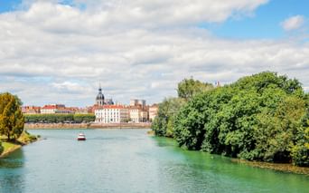 Blick auf die Saône in Chalon-sur-Saône mit grünen Bäumen am Ufer, einem kleinen Boot auf dem Wasser und historischen Gebäuden unter blauem Himmel.
