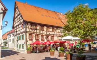 Traditional half-timbered building with orange tile roof and outdoor café with red umbrellas in Fridingen old town square under blue sky.