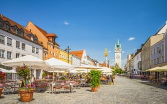 Kopfsteinpflasterplatz in Straubing mit Straßencafés, bunten historischen Gebäuden und dem Stadtturm mit grüner Spitze unter blauem Himmel.