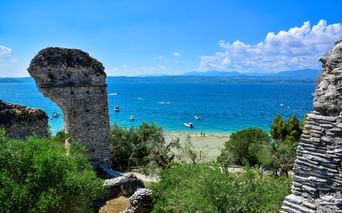 Antike Steinturmruinen in Sirmione mit türkisfarbenem Gardasee, Booten, Strand und Bergen unter blauem Himmel.