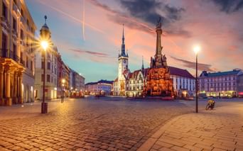 Olmützer Marktplatz in der Dämmerung mit beleuchteter Dreifaltigkeitssäule und Rathausturm. Kopfsteinpflaster und historische Gebäude unter buntem Himmel.