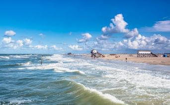 Breiter Sandstrand in St. Peter-Ording mit rollenden Wellen, Pfahlbauten in der Ferne und dramatischen Wolken am blauen Himmel.