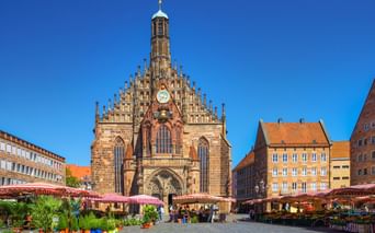 Frauenkirche church at Nuremberg's Hauptmarkt square with market stalls. Gothic facade with ornate gables, clock, and tower against blue sky.