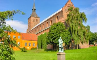 Bronzestatue von H.C. Andersen auf grüner Wiese in Odense, mit rotem Backsteinkirchturm und gelbem historischen Gebäude im Hintergrund.