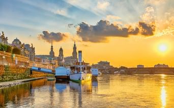Sunset view of Dresden's Brühlsche Terrasse with boats moored on the Elbe River. Historic buildings and church towers visible against golden sky.