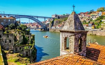 Blick auf die Ponte Dom Luís I in Porto über dem Douro, mit Kirchturm im Vordergrund und bunten Häusern am Hang.