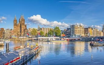 Blick auf Amsterdamer Hafen mit den Zwillingstürmen der St. Nikolaus-Basilika, historischen Gebäuden, Anlegestellen und Booten auf ruhigem Wasser.