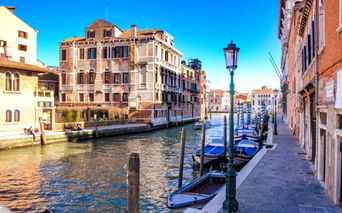 Venezianischer Kanal mit vertäuten Gondeln entlang eines Gehwegs. Historische Gebäude säumen den Wasserweg unter blauem Himmel. Traditionelle Laternen stehen entlang der Promenade.