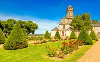 Abteikirche mit achteckigem Turm in Saint-Florent-le-Vieil, umgeben von formalen Gärten mit kegelförmigen Hecken, bunten Blumenbeeten und Bäumen.