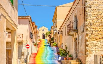 Bunte Regenbogentreppe in einer engen Gasse in Arzachena, Sardinien, gesäumt von traditionellen beigen Gebäuden unter blauem Himmel.